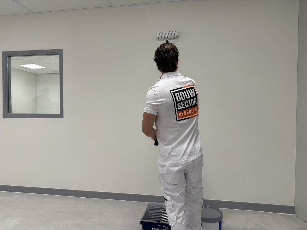 Schilder in witte werkkleding rolt een lichte wand naast een binnenraam met grijs kozijn
