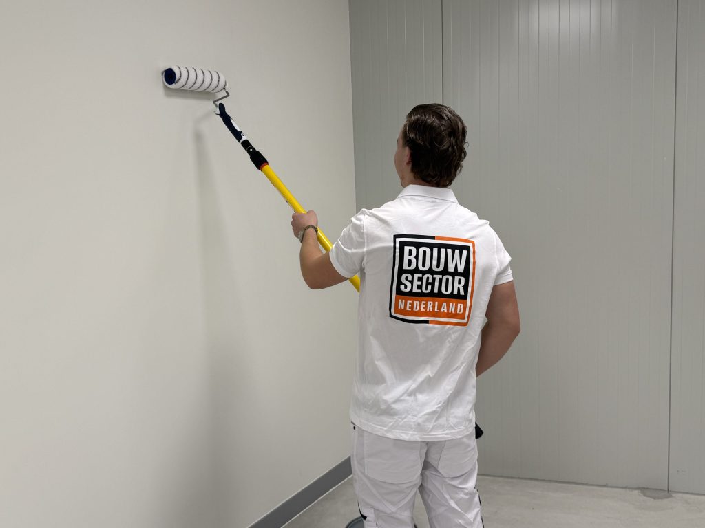Schilder van Bouwsector Nederland saust met een lange roller een witte binnenmuur in een lege ruimte