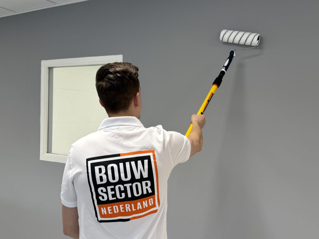 Schilder met Bouwsector-logo op de rug saust een grijze wand naast een wit geschilderd raamkozijn