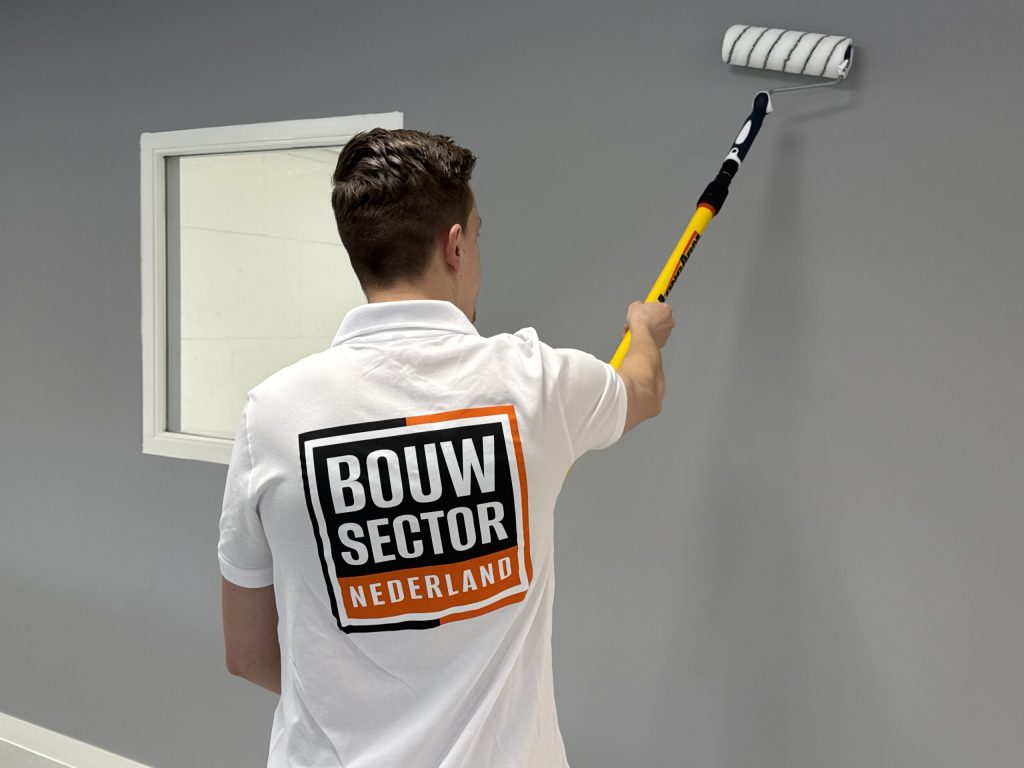 Schilder rolt met een verlengsteel een grijze muur naast een raam met witte omlijsting
