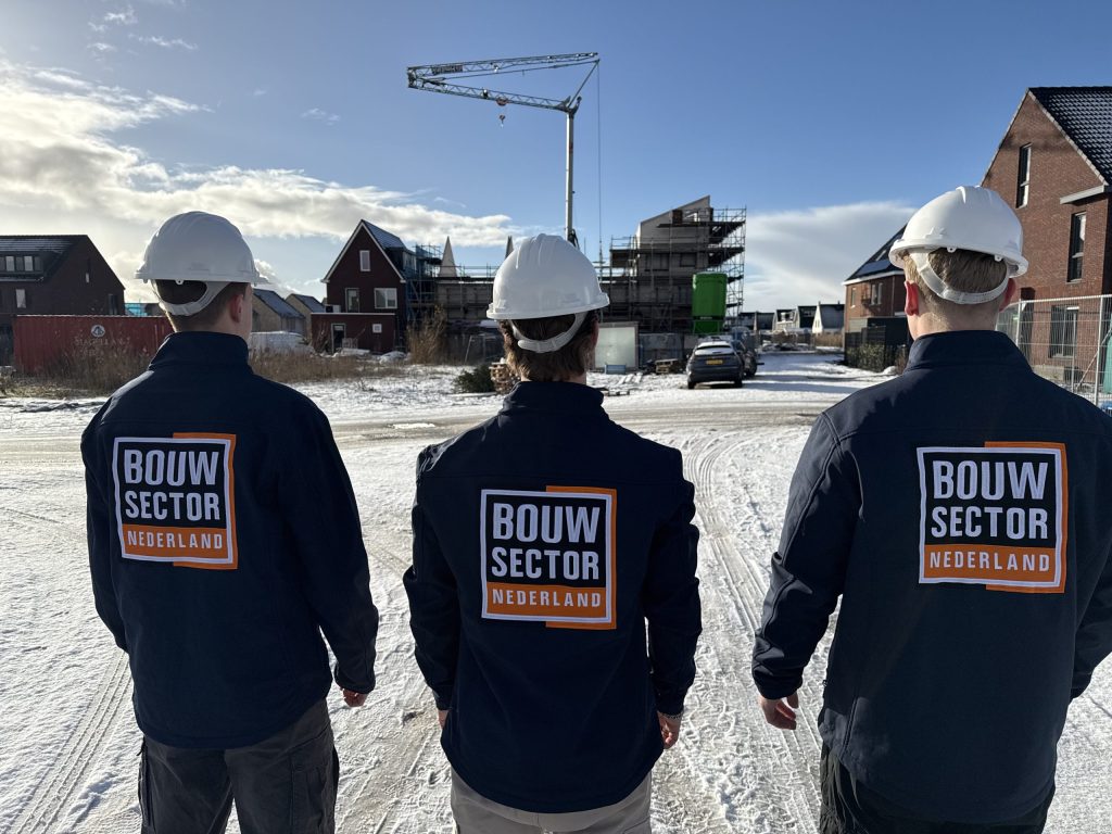 Drie vakmensen van Bouwsector Nederland met witte helm kijken naar bakstenen woningen en een bouwkraan in de sneeuw