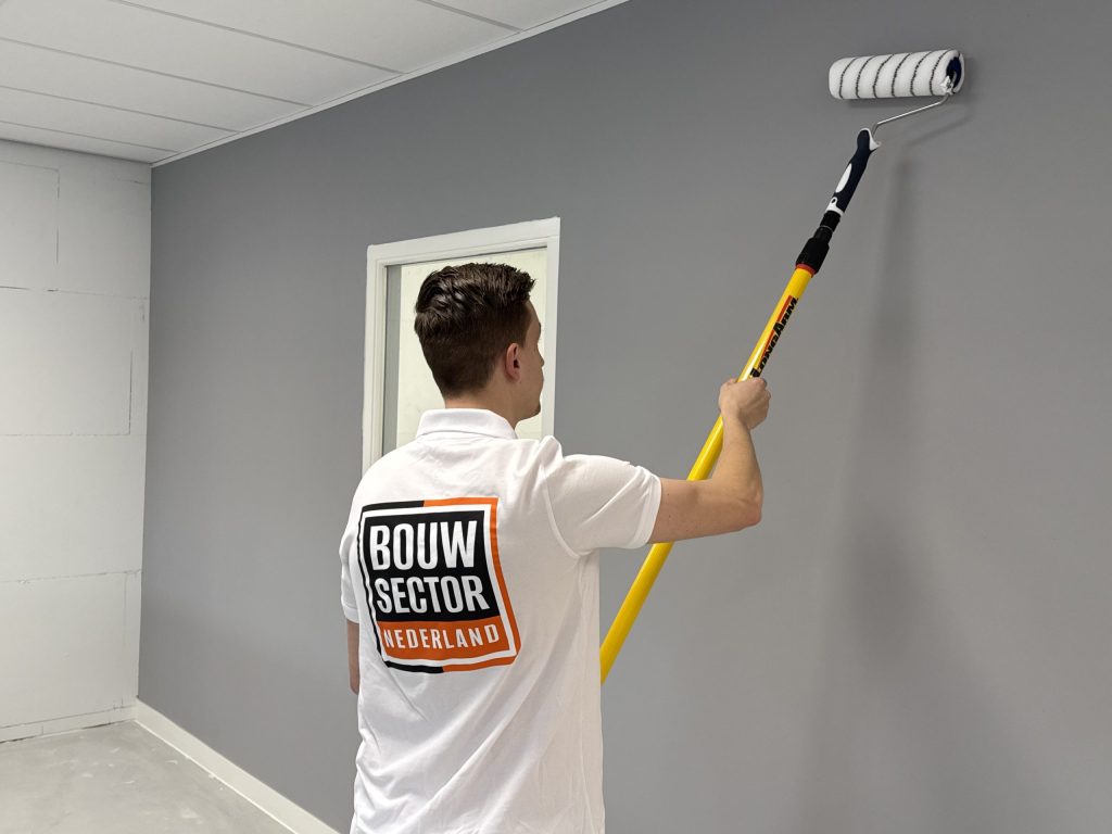 Schilder rolt een grijze muur onder een plafond van systeemplaten, naast een deuropening